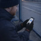Man at a bus stop uses his smartphone wearing Acrylic Blend Tech Gloves on a cold day.