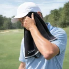 Golfer in white cap wipes face with a Titleist Players Towel on a sunny golf course.