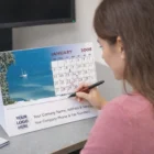 Woman writing on a 12 Page Desk Calendar at an office desk with a computer monitor.