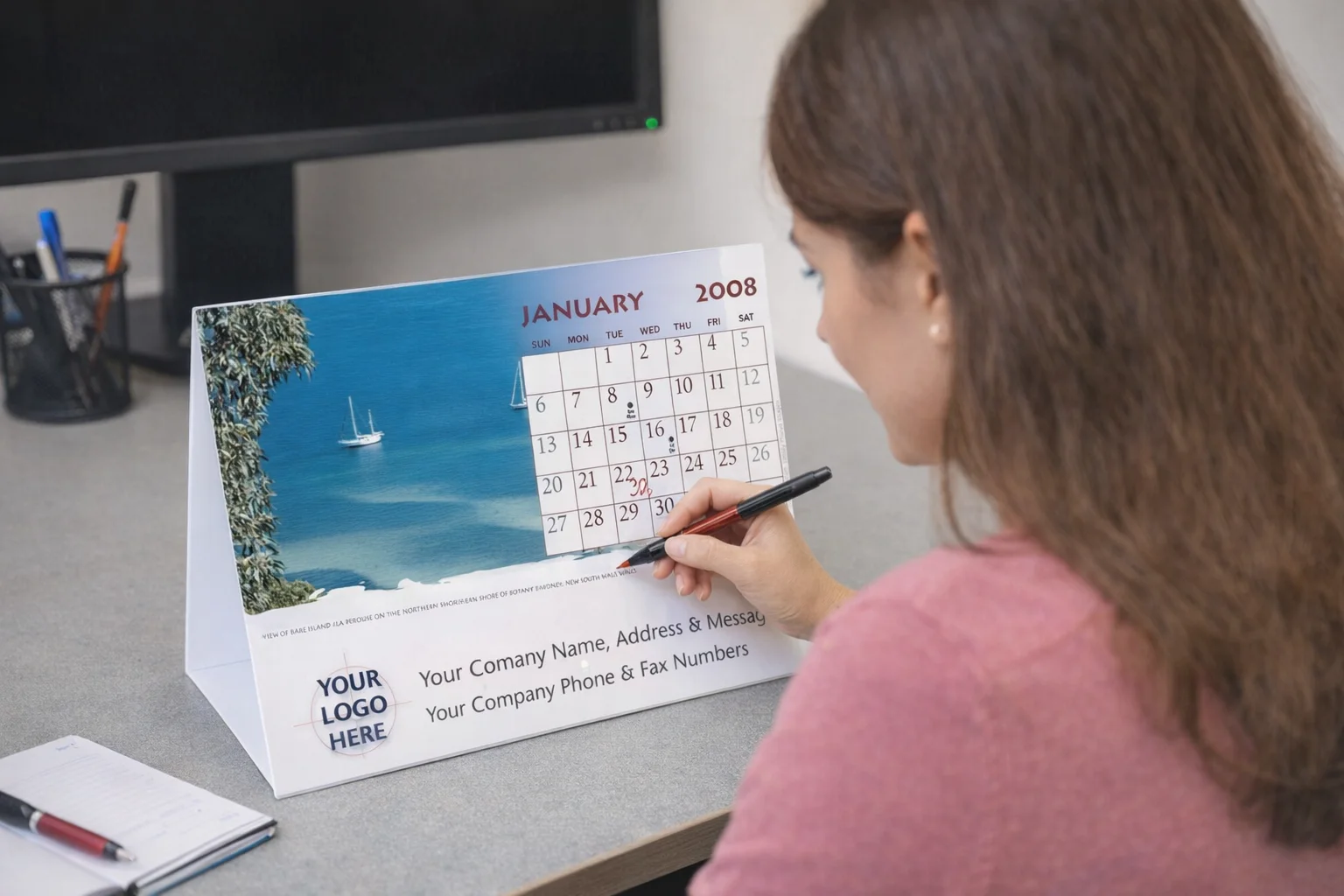 Woman writing on a 12 Page Desk Calendar at an office desk with a computer monitor.