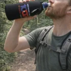 Hiker drinks from Water Bottle Cooler With Carabiners (Sublimation) on a forest trail.