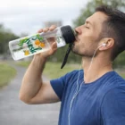 Man in blue shirt drinks from 700Ml Light Up Drink Bottle while wearing earphones outdoors.