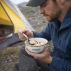 Man enjoys oatmeal outdoors near a yellow tent using Enamel Finish Bowls For Camping.