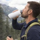 Man outdoors with a backpack, drinking from a Dakota Vacuum Bottle, mountains in the background.