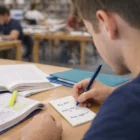 Person writing notes with Caspian Office Note Pads 90mm x 160mm, an open book, and a highlighter.