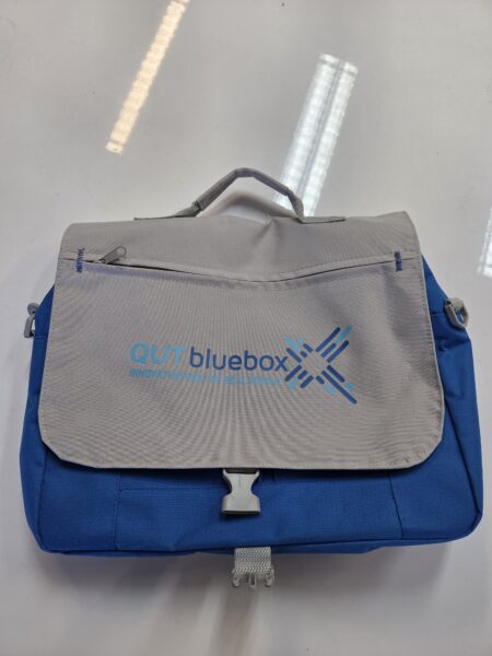 Blue and gray QUT bluebox branded merchandise bag with handle and front flap on a white surface.