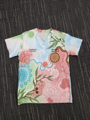 A colorful, age-inclusive scrub top with Aboriginal art patterns laid flat on a dark textured floor.