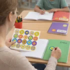 A teacher places a 44mm glossy circle sticker in a notebook at a classroom table.