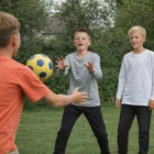 Three boys in Youth Staple L/S Tees play catch with a yellow and blue soccer ball on grass.