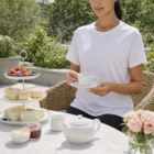 Woman in Wo's Maple Curve Tee enjoys tea and pastries at a flower-filled garden table.