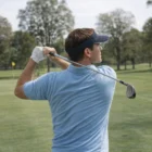 Man wearing Edge Visors swinging golf club on green course with trees in the background.
