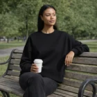 Woman in Wo's Heavy Crewss sits on a bench with coffee, trees in the background.