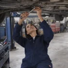 A mechanic in Wo's Canvas Coveralls works under a lifted car in an auto repair shop.