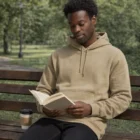 Man in a beige Stencil Hoods hoodie reading on a park bench with coffee.