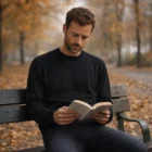A man in a Mens Knit Crewss sits on a park bench reading among autumn trees and fallen leaves.