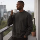 Man wearing Heavy Crewss sweatshirt holding a white mug on a balcony with city view.