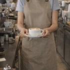 Barista wearing Canvas Bib Aprons holds a cappuccino in a coffee shop.