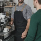 Barista in Village Denim Bib Apron smiles while making coffee at a cozy coffee shop counter.