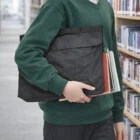 Student in green sweater with books and a Satchel Bag walks through a library aisle.