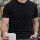 Man wears Men's Combed Cotton Tee, sits outdoors with a mug at a wooden table.