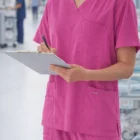 Person in Carevibe Unisex Pink V-Neck Scrub Top writing on a clipboard in a hospital hallway.