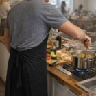 Man in gray shirt wearing Aprons - Long, cooking at a stove with vegetables in the kitchen.
