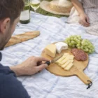 A person slices cheese on a Lynx Serving Board, with grapes, salami, bread, and wine nearby.