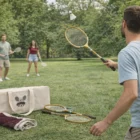 Three play with Sporty Backyard Badminton Sets; rackets and bag rest nearby in the park.