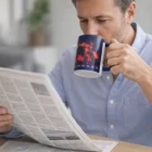 Man in blue shirt drinks from a Colour Max Can Mug while reading a newspaper at the table.