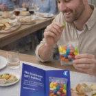 Smiling man holds a Gift Card With 50G Aussie Jelly Bean Bags and a brochure at a dining table.