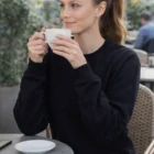 Woman wearing a Fleecy Sweat Crewss sits at an outdoor café, holding a cup and smiling.