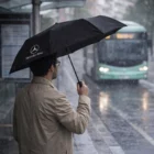Man holding a Poppins Umbrella at a bus stop as a green bus arrives in heavy rain.