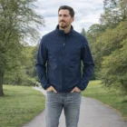 Man in Mens Stormtech Ecoguard Softshell Jacket and jeans stands on a park path with trees.