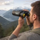 Man uses 10 X 50 Binoculars to view mountains and a lake from a scenic overlook.