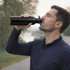 Man in a navy jacket drinks from The Athlete 800Ml Metal Bottle outdoors on a path.