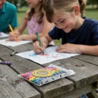 Smiling child colors at picnic table with Louvelle Crayons in PVC zipper pouches beside them.