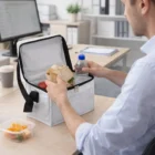 Man eating lunch at his desk with a Lunch Chiller Bag With Logo Print, sandwich, and water bottle.
