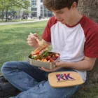 A young man eats a sandwich from his Miranda Lunch Box while sitting on the grass.