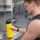 Wearing earbuds, man fills Amara 750ml Bottle with flip straw lid at gym water station.