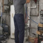 A man in Lads Utility Cargo Pants repairs pipes with tools at his side in an industrial room.
