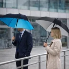 Two people with Marques 23" Square Automatic Umbrellas stand in the rain by a building.