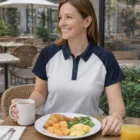 Woman in Ladies Harmony Polo smiles at outdoor cafe with breakfast, coffee, and juice.