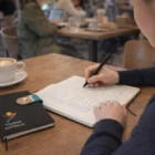 Someone writes in a Bio Plus Rpet A5 Notebook at a café with coffee, phone, and booklet.
