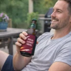Smiling man holds a beer outdoors with a Stubby Cooler Wraps sleeve.