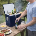 Person holding a beer by a Bambin Vintage Cooler Box on an outdoor picnic table with food and drinks.