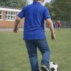 Kid wearing Conference Polo Shirts Kids and jeans kicks a soccer ball on grass by the school.