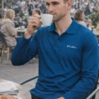 Man in Mens Truedry Custom Logo Polos Long Sleeve drinks coffee at an outdoor café.