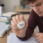 Person holding a Stress Balls Golf while writing at a desk with books.