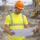 Man in Hi Vis Mens T-Shirts With Custom Branding reviews blueprints at construction site.