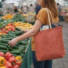 Person at a market holds zucchini with a Skyler Herringbone Pattern Jute Tote Bag, 29L.